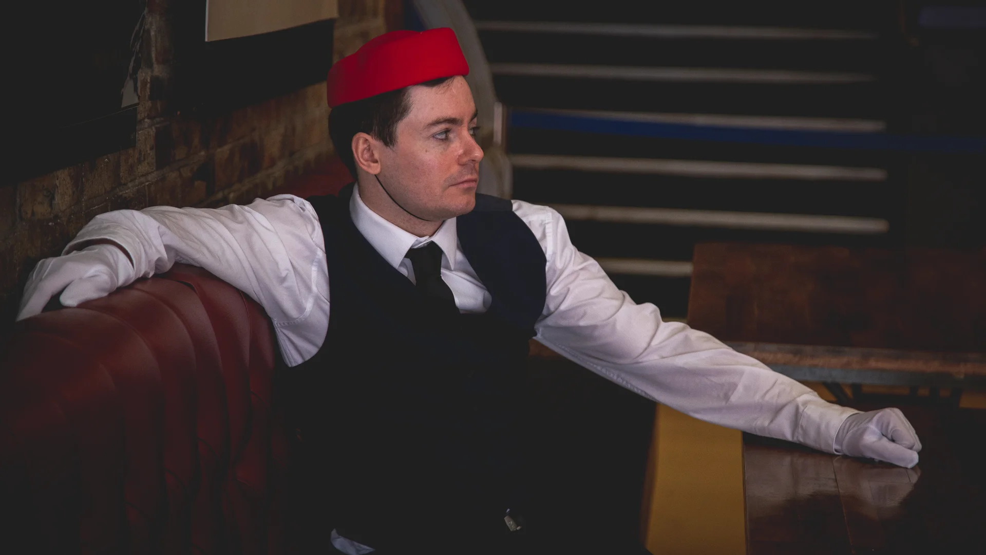 Luke Hunter wearing a red hat, seated at a table in Tickets Please