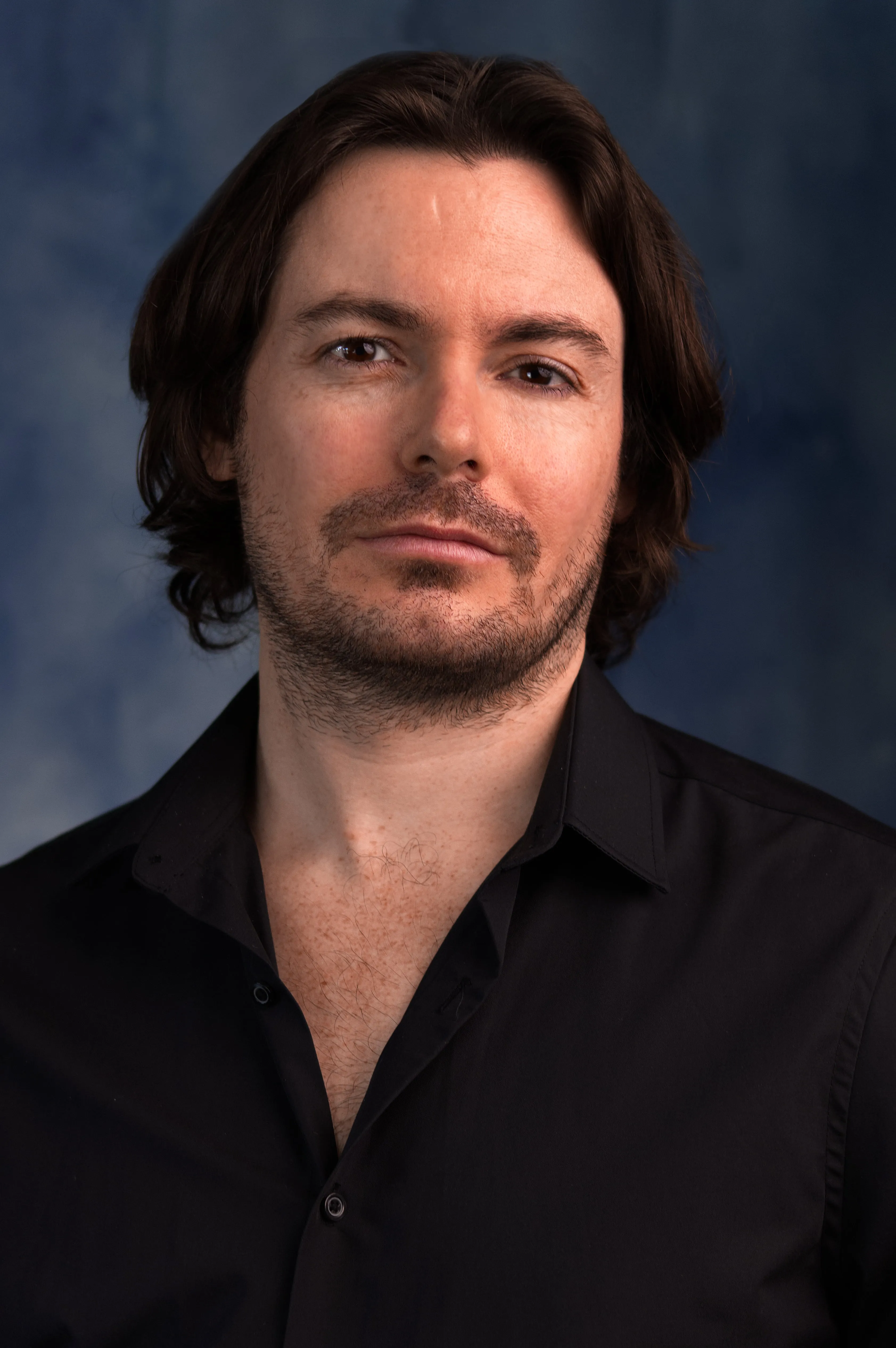 Luke Hunter in a black open-collar shirt against a blue-grey background